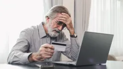 sivovlasý muž si drží hlavu, v druhej ruke platobnú kartu, pred ním laptop