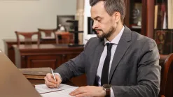 muž v sivom obleku s kravatou podpisuje perom dokumenty za stolom v kancelárii s dreveným nábytkom