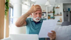 Muž si dal ruku na hlavu, v druhej drží list, pred ním na stole otvorený notebook