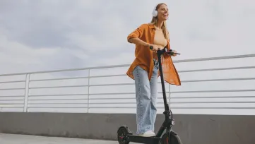 usmiata žena so slúchadlami na elektrickej kolobežke na promenáde pri zábradlí
