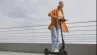 usmiata žena so slúchadlami na elektrickej kolobežke na promenáde pri zábradlí
