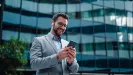 Muž v saku s mobilom stojí pred presklenou kancelárskou budovou