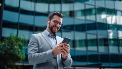 Muž v saku s mobilom stojí pred presklenou kancelárskou budovou