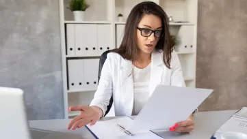 Žena v kancelárii prekvapene číta dokument, na stole doklady a laptop