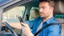 Muž v modrej košeli drží počas jazdy autom v pravej ruke mobilný telefón