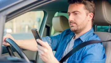 Muž v modrej košeli drží počas jazdy autom v pravej ruke mobilný telefón
