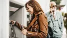 Usmiata žena s mobilom pri bankomate, v pozadí telefonujúci muž