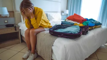 Žena sedí na hotelovej posteli s hlavou v dlaniach, vedľa otvorený kufor