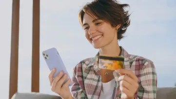 Usmiata mladá žena vonku drží mobil a platobnú kartu