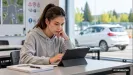 mladá žena v sivej mikine v učebni autoškoly robí test z teórie na tablete, za oknom stojí biele auto
