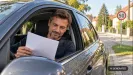 muž za volantom zaparkovaného auta číta papier, v pozadí ulica v obci a značka 50 km/h